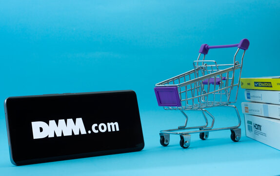 Dhaka, Bangladesh- 03 Oct 2025: Smartphone with Dmm.com logo next to a small shopping cart and boxes against a blue background.