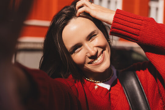 Close up selfie portrait of pretty girl with long dark hair on street at city urban background. She wear red sweater, black bag. Smiling girl, happy, positive emotions. Woman fix her hair, blink eye - Powered by Adobe