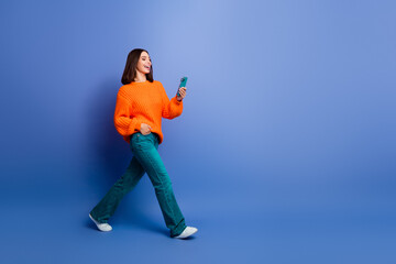 Young Woman in an Orange Sweater Walking and Smiling Against a Blue Background With a Smartphone in Hand