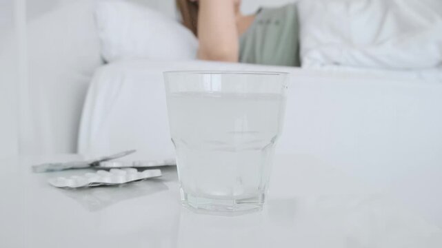 Sick woman lying in bed reaching for glass of effervescent cold medicine on bedside table, suffering from headache and high temperature. Female with viral disease. Health care and recovery concept