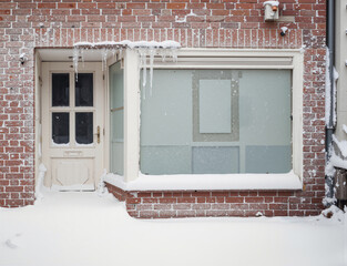 Closed shop entrance in winter with a vintage wooden door and frosted windows, surrounded by red brick walls. Snow and icicles cover the facade, creating a cold, deserted and atmospheric urban winter 