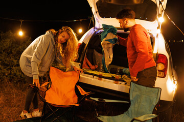 Couple setting up chairs for a cozy nighttime camping experience under string lights next to their car