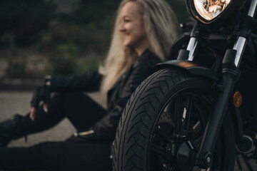 Smiling woman in black leather jacket sitting on the ground near a motorcycle, enjoying freedom and...