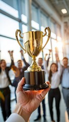 Hand holding golden trophy in front of cheering business team celebrating success and achievement in office.