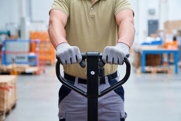 Warehouse worker wearing gloves operating pallet jack for manual handling and transportation of goods.
