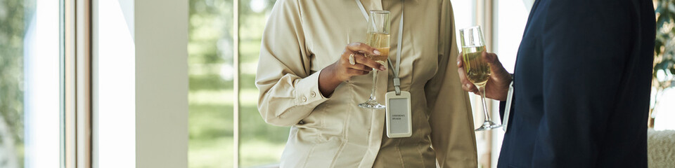 Caucasian middle aged woman and Black middle aged man holding champagne glasses and talking near window during business event, both wearing formal clothing and conference badges