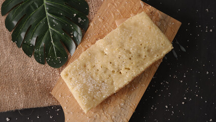 Rectangular shortbread cookie on a wooden board next to a tropical leaf, sprinkled with powdered sugar on a dark surface.