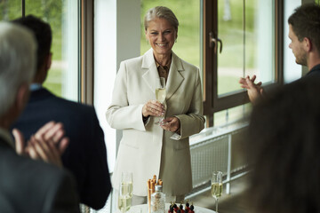 Senior Caucasian woman smiling while holding champagne glass, standing near table with desserts, surrounded by applauding adults in bright room with large windows during celebration