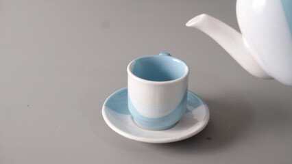 A white and blue teapot pours liquid into a matching cup sitting on a saucer against a gray background.