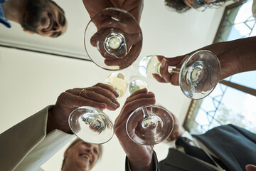 Group of diverse adults clinking wine glasses during celebration, multiethnic men and women smiling and toasting, hands and faces visible from low angle, indoor gathering