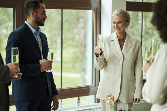Middle aged Caucasian woman smiling and holding champagne glass, while standing with young adult Caucasian man and diverse colleagues during business celebration near large windows