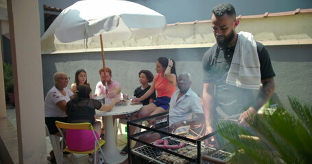 Diverse group enjoying backyard barbecue together, featuring a man grilling and others gathered at a table under an umbrella, emphasizing joyful social interaction