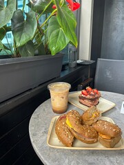 Three sesame bread rolls, iced coffee, and chocolate cake with strawberries are served on a small round table next to a plant on an outdoor balcony.