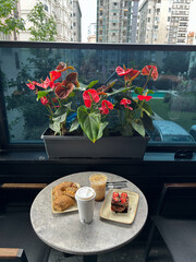 A round table with a paper coffee cup, iced coffee, two sesame pastries, and a chocolate strawberry cake is placed on a balcony with city buildings.