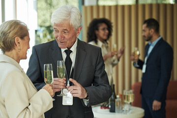 Senior Caucasian man talking with senior Caucasian woman while holding champagne glasses at business networking event, diverse group of young adults conversing in background