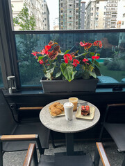 Outdoor caf table with two coffee cups, pastries, and a dessert slice, with red flowers and high-rise buildings in the background.