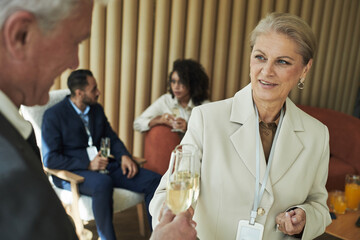 Senior Caucasian woman holding champagne glass and smiling at senior Caucasian man during business networking event, multiethnic colleagues sitting and talking in background