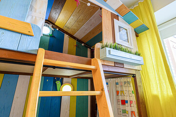 A colorful, playful children's bedroom with a unique wooden loft bed resembling a treehouse. The bed is accessed by a wooden ladder. Yellow curtains add warmth to the room