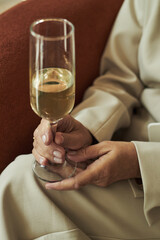 Senior Caucasian woman holding champagne glass with both hands, sitting on chair, showing manicured nails and relaxed posture, partial body visible, no face in frame
