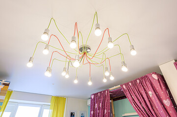 A modern ceiling light fixture with multiple exposed bulbs hanging from colorful (green, red) wires, suspended from a chrome base. Located in a playful room with bright curtains and a canopy bed
