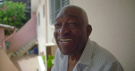 One Elderly Black man sitting outdoors on porch, laughing warmly, exuding joy and positivity, tropical residential surroundings with pink walls and greenery in background