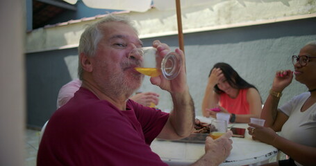 Elderly man smiling and taking a sip of beer from a glass at a backyard barbecue, embodying joy and connection during a lively gathering with family and friends
