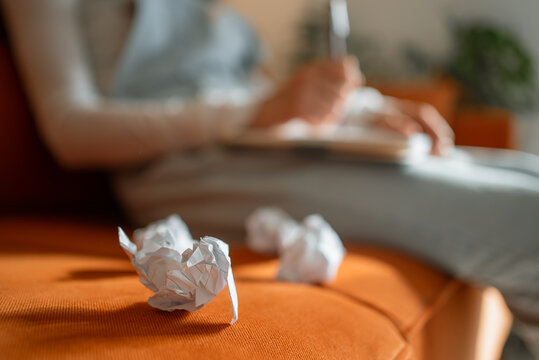 Crumpled paper balls lie on an orange sofa, symbolizing a creative crisis while a writer works on a notebook in the background, highlighting the challenges of the creative process