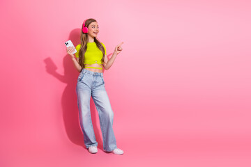 Cheerful young woman with headphones holding smartphone pointing to side on pink backdrop in casual stylish outfit