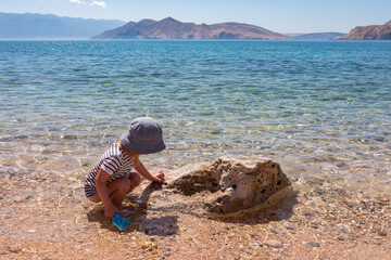 A curious little girl in a sun hat crouches by a rock on a pebble beach in Baska, Croatia, playing...