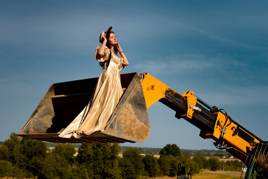 A beautiful woman in an opulent golden dress sits regally in a backhoe bucket against a clear blue sky and lush green landscape, creating a surreal and striking juxtaposition.