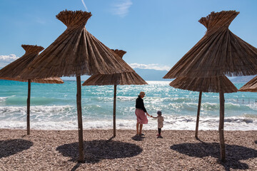A mother and her daughter stand together on a scenic pebble beach with straw umbrellas, enjoying the beautiful turquoise Adriatic Sea view on a summer day in Baška, Krk, Croatia.
