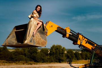 A striking young woman in a golden dress sits gracefully on a construction tractor bucket in a...