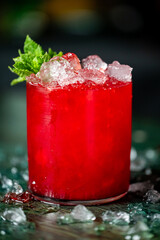 Red cocktail with crushed ice, mint sprig, and berry garnish in a clear glass. Refreshing summer drink on ice-covered surface. Close-up, vibrant colors, top view.