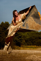 Fototapeta premium Elegant woman in golden dress confidently poses inside an excavator bucket in field, creating a striking contrast between glamour and rugged machinery, radiating beauty and empowerment in nature.