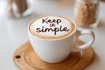 Coffee cup with foam art displaying the words keep it simple, resting on a wooden coaster, surrounded by a cozy cafe atmosphere, inviting relaxation and enjoyment