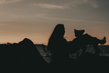 Silhouette of a woman and her small dog at sunset. A woman holds her Yorkshire terrier doggy up as they enjoy the sunset over the water