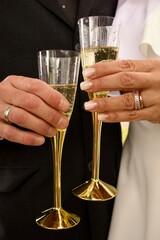 Two hands with wedding rings holding champagne glasses with golden stems, symbolizing celebration, wedding, and a romantic toast.