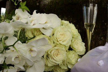  A white wedding bouquet of roses and orchids rests beside a glass of champagne and satin fabric, creating an atmosphere of celebration, elegance, and romance.