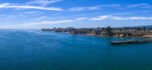 Aerial view of Capitola, California, showing a wooden pier, rugged cliffs, a calm blue ocean, coastal buildings, and a clear sky with scattered clouds.