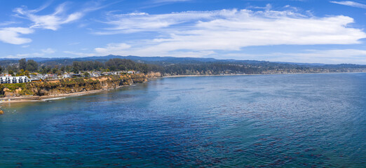 Aerial view of Capitola, California, showing rugged cliffs with houses, calm Pacific waters, rolling hills, and a clear sky with scattered clouds.