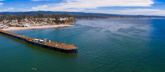 Fototapeta premium Aerial view of Capitola Beach in California with a wooden pier, colorful coastal buildings, rolling hills, trees, and a clear sky with scattered clouds.