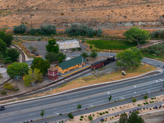 Aerial view of a yellow train station with green roof, red signal tower, vintage locomotive, parking area, trees, and desert landscape in Carson City.