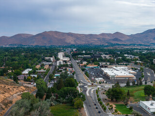 Aerial view of Carson City, Nevada, showing roads, commercial and residential areas, green spaces, and rugged mountains under a partly cloudy sky.