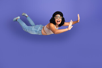 Joyful young woman pretending to fly in casual outfit on a vibrant purple background, expressing...