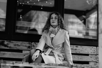 Young woman with curly hair wearing a stylish coat, posing confidently against a wooden structure, showcasing modern fashion and elegance in a monochrome setting