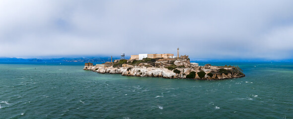 Aerial view of Alcatraz Island in San Francisco Bay, featuring the historic penitentiary, rocky shoreline, and surrounding blue green waters under a cloudy sky.