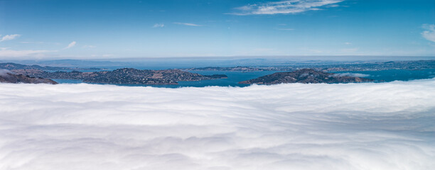 Aerial perspective of the San Francisco Bay area, featuring white clouds in the foreground, hilly terrain, coastal areas, and a clear blue sky.