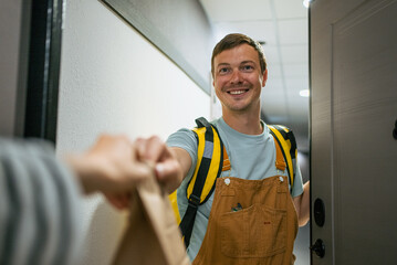 Friendly delivery man wearing overalls and a backpack is handing over a brown paper bag with a meal to a customer at their doorstep, representing efficient and modern service