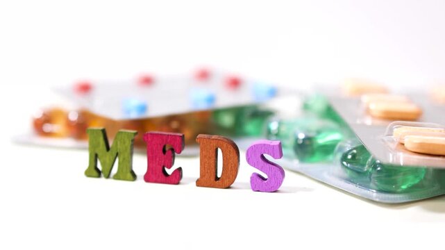 Rotating view of colorful MEDS wooden text in front of medicine pills on white background.