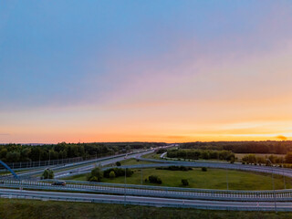 An aerial view of a modern highway at dusk. The sun sets in the background, casting warm hues across the sky. Lush greenery flanks the road, creating a serene landscape.